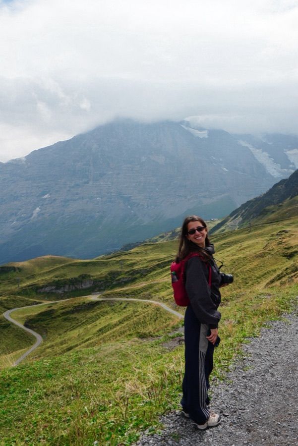 Danielle smiling on a gravel trail, with a winding mountain road and a cloud-capped peak behind her